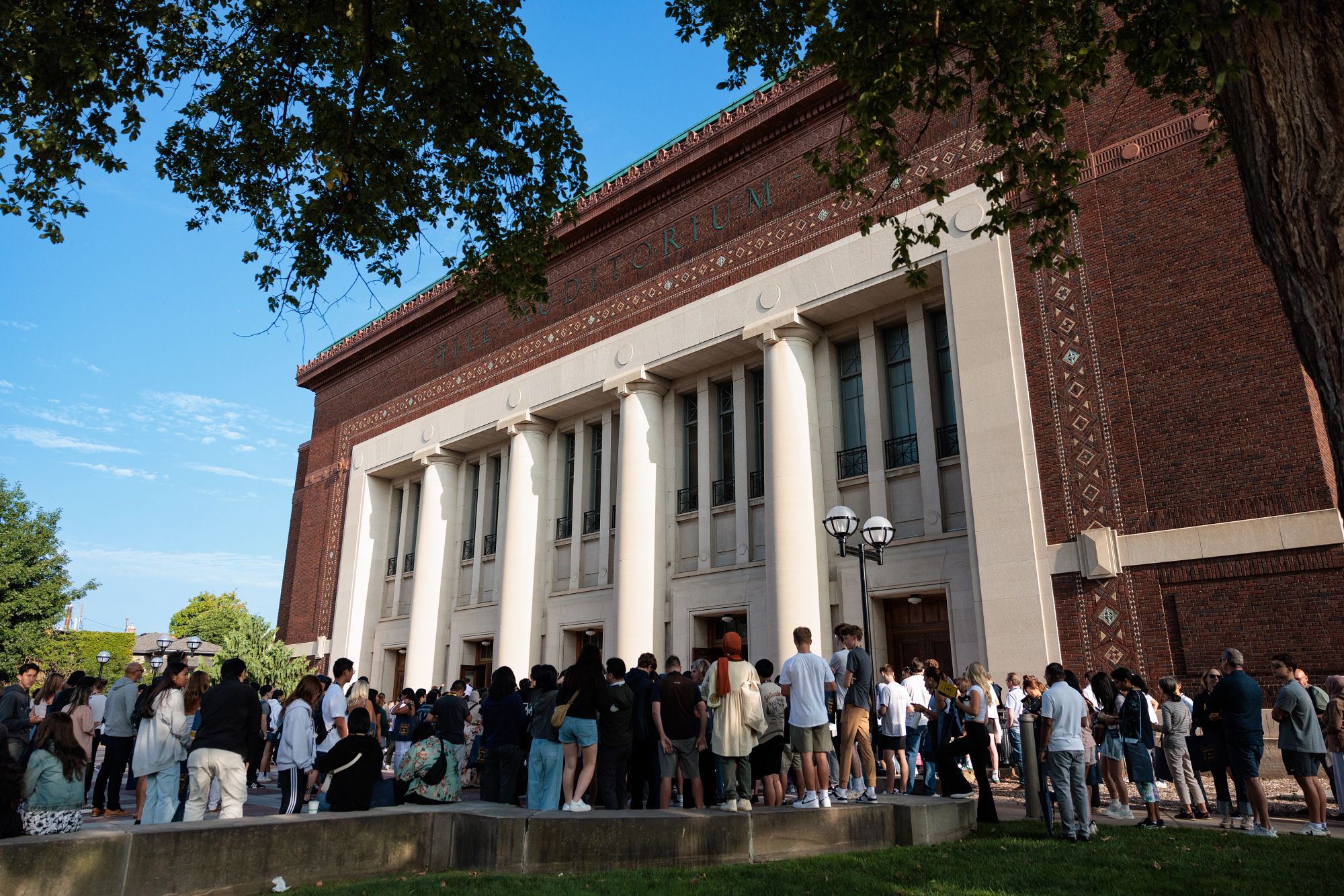 A large group gathers outside of Hill Auditorium on a clear summer morning.