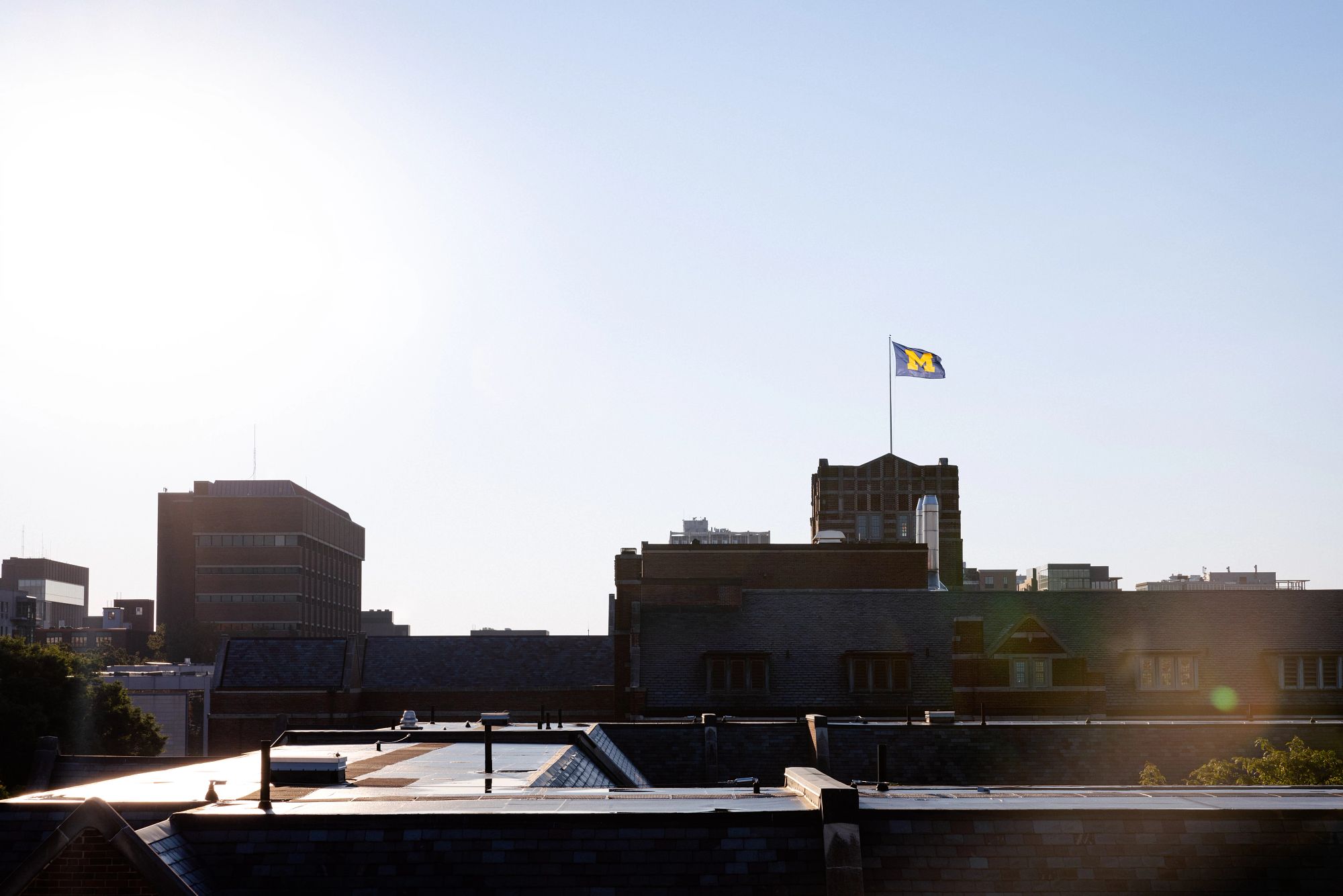 The flag on top of the Michigan Union peeks out from behind the rooftops of West Quad against a bright sky. Puddles reflect the bright morning sunlight on the West Quad roofs