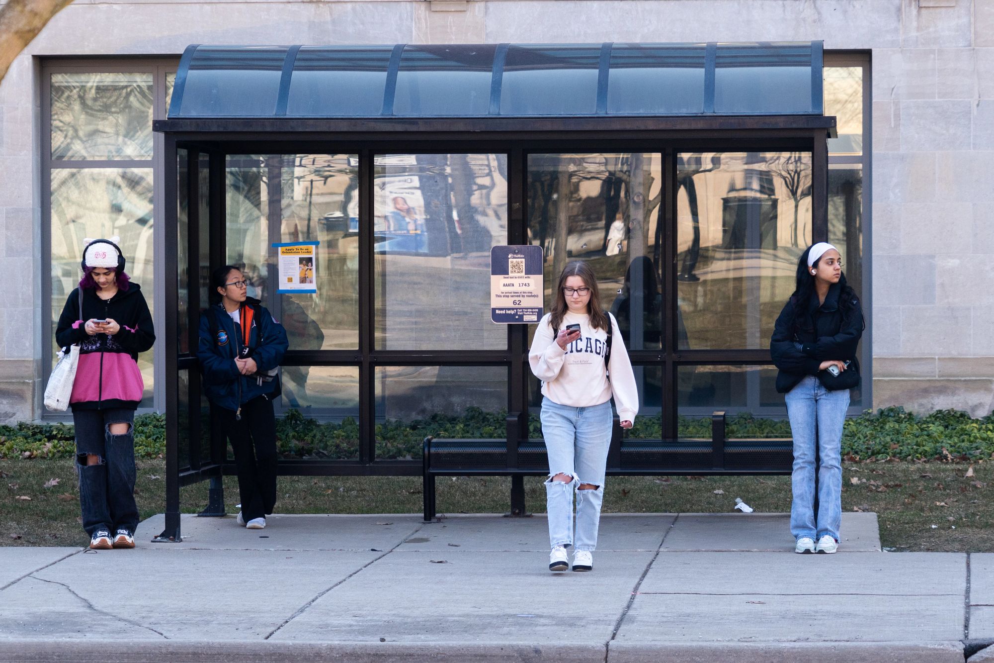 Four students stand in front of a bus stop near Ingalls Mall. Two look at their phones, while two lean against the bus stop enclosure.