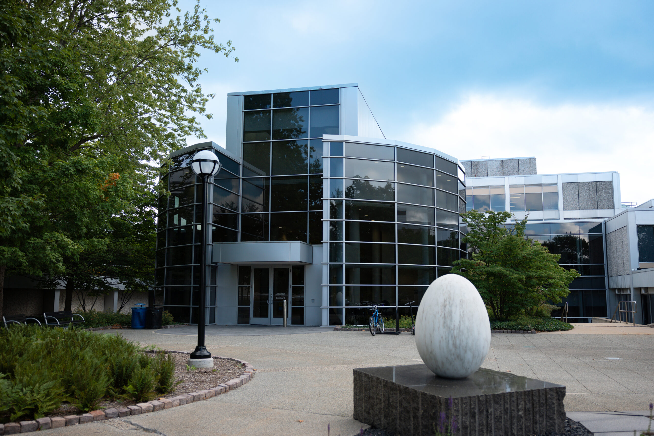 Building 16 of the North Campus Research Complex is made up of two semi-circular structures with large walls made up of curved black windows. A more traditional rectangular building sits in the middle of both curved structures with an awning and doorway at its base. Nearby on the sidewalk is a large egg-shaped sculpture called “Liberation”